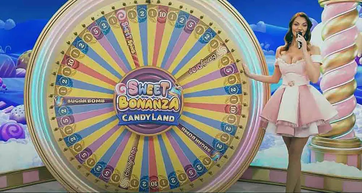 “A game show host in a pink dress stands beside the large Sweet Bonanza CandyLand prize wheel on a bright, candy-themed stage with pastel clouds and colorful lighting.”