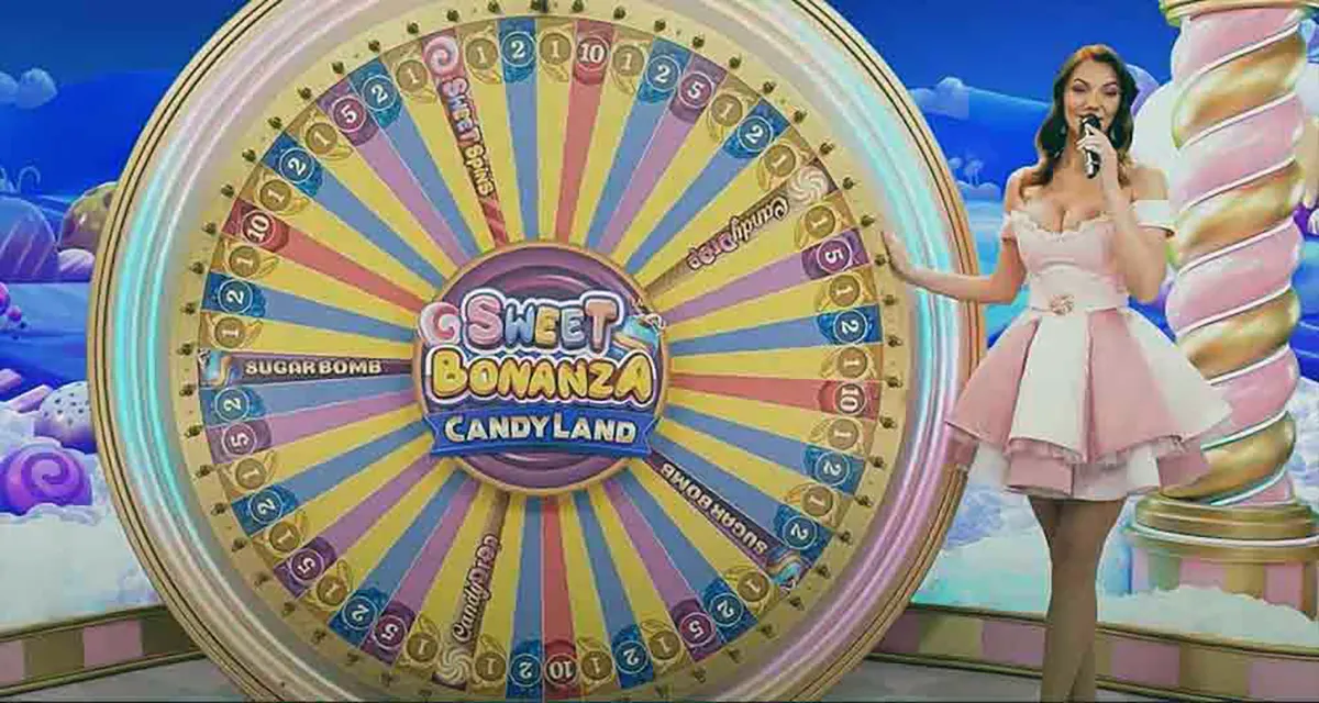 “A game show host in a pink dress stands beside the large Sweet Bonanza CandyLand prize wheel on a bright, candy-themed stage with pastel clouds and colorful lighting.”