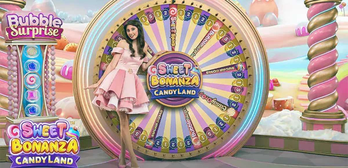 “A presenter in a pink dress poses beside the large Sweet Bonanza CandyLand bonus wheel on a colorful candy-themed game show set with pastel clouds and confectionery decorations.”